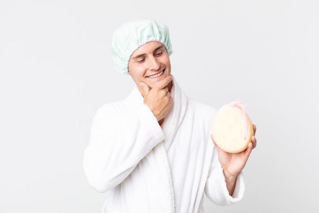 Young handsome man smiling with a happy, confident expression with hand on chin with bathrobe, shower cap and a sponge