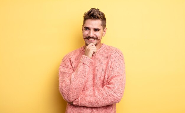 Young handsome man smiling with a happy, confident expression with hand on chin, wondering and looking to the side. strange moustache concept