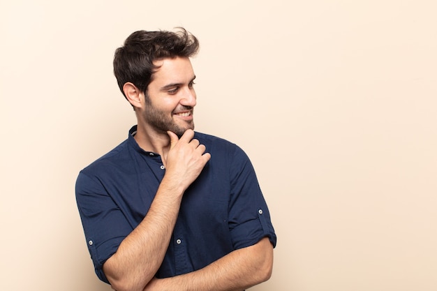 Young handsome man smiling with a happy, confident expression with hand on chin, wondering and looking to the side