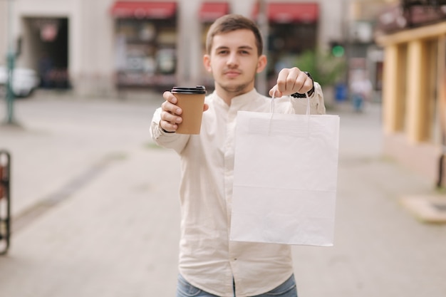 若くてハンサムな男が街に立って、パッケージとコーヒーを保持します