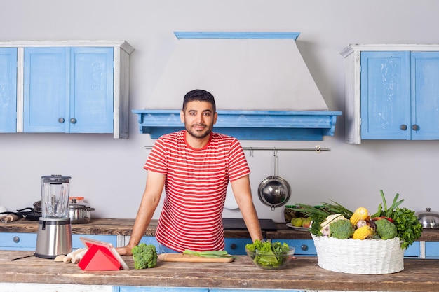 Young handsome man standing in kitchen with modern furniture looking at camera with smile basket of fresh green vegetables and blender on table vegetarian food vegan nutrition healthy diet