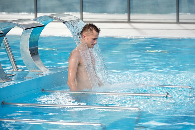 Young handsome man standing in relaxing spa waterfall in blue transparent water of swimming pool and enjoying time at resort