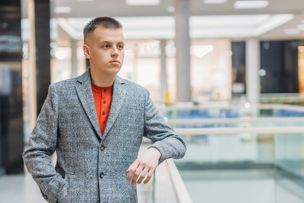 Photo young handsome man stands in a light coat in the building with his hand on the fence.