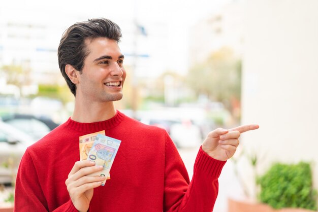 Young handsome man taking a lot of money at outdoors pointing to the side to present a product