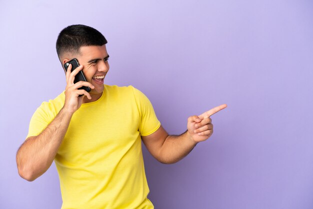 横に指を指し、製品を提示する孤立した紫色の背景の上に携帯電話を使用して若いハンサムな男