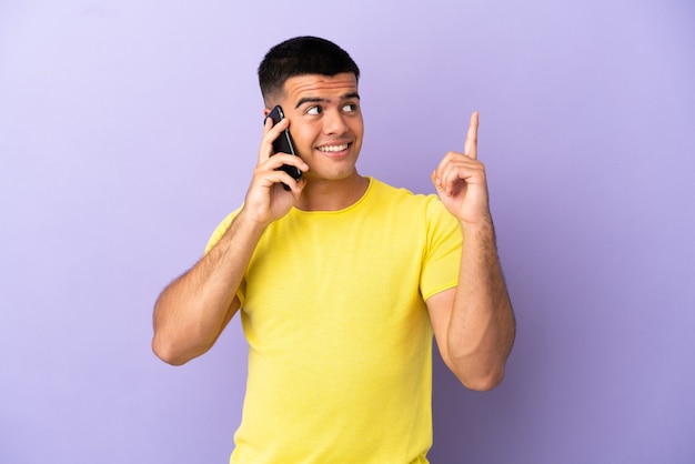 Young handsome man using mobile phone over isolated purple background pointing up a great idea