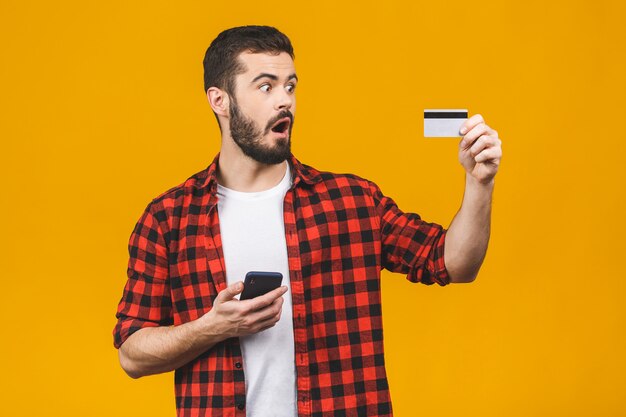 Il giovane uomo bello che per mezzo dello smartphone ha isolato spaventato nella scossa con un fronte di sorpresa, impaurito ed eccitato con espressione di paura con la carta di credito, facente la spesa.