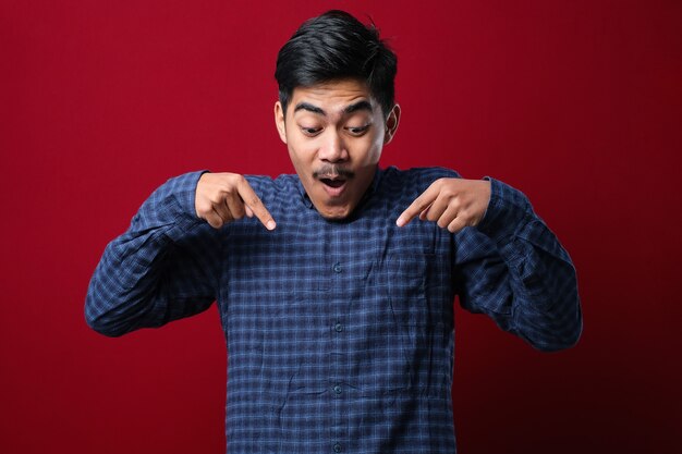 Young handsome man wearing casual shirt standing over red background Pointing down with fingers showing advertisement, surprised face and open mouth