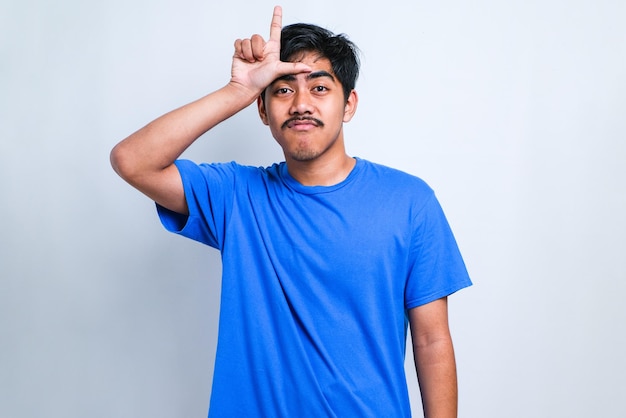 Young handsome man wearing casual shirt standing over white\
isolated background making fun of people with fingers on forehead\
doing loser gesture mocking and insulting.