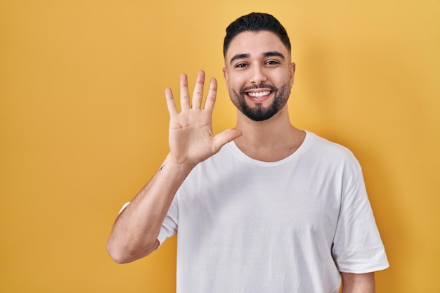 Young handsome man wearing casual t shirt over yellow background showing and pointing up with fingers number five while smiling confident and happy