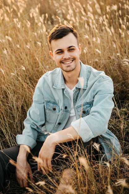 Giovane uomo bello, indossa una giacca di jeans, seduto in un campo, sorridente.