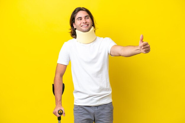 Young handsome man wearing neck brace and crutches isolated on yellow background giving a thumbs up gesture