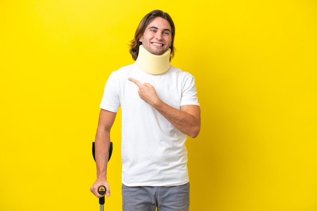 製品を提示する側を指している黄色の背景に分離された首ブレースと松葉杖を身に着けている若いハンサムな男