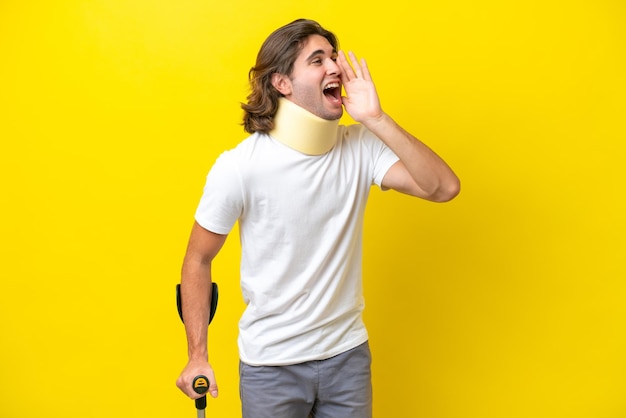 Young handsome man wearing neck brace and crutches isolated on yellow background shouting with mouth wide open to the side