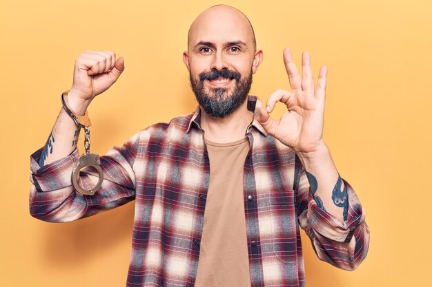 Young handsome man wearing prisoner handcuffs doing ok sign with fingers, smiling friendly gesturing excellent symbol