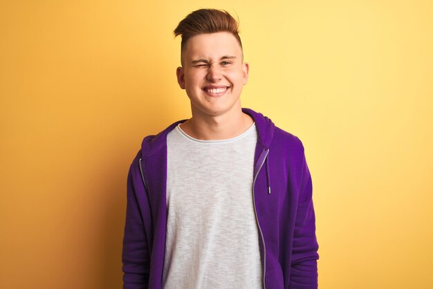 Young handsome man wearing purple sweatshirt standing over isolated yellow background winking looking at the camera with sexy expression cheerful and happy face