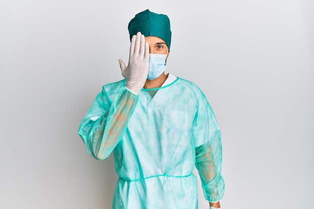 Young handsome man wearing surgeon uniform and medical mask covering one eye with hand, confident smile on face and surprise emotion.