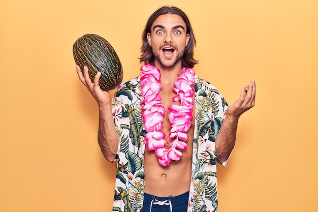 Young handsome man wearing swimwear and hawaiian lei holding melon celebrating achievement with happy smile and winner expression with raised hand