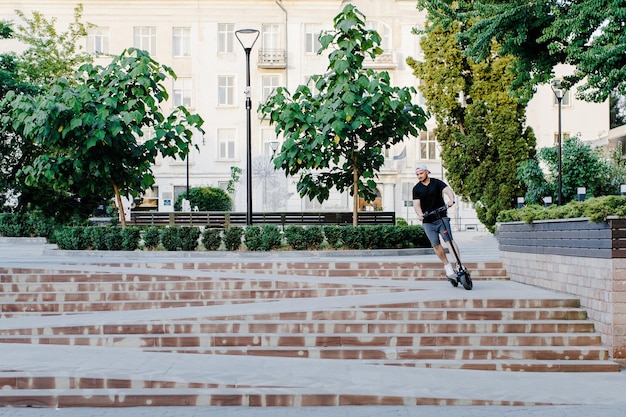 Young handsome man in white baseball cap riding on scooter to work along the street in the city