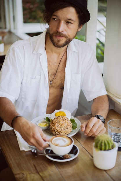 Young handsome man in a white shirt open, having breakfast in a cafe with a vegetarian burger, drinking coffee, lifestyle in a tropical island, life in Bali.