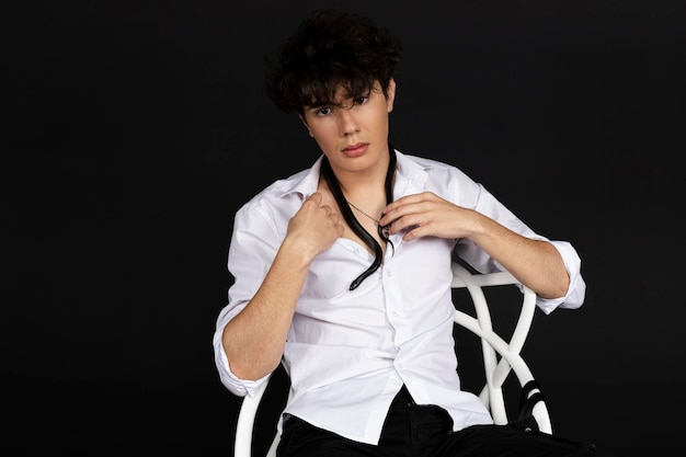 Young handsome man in a white shirt sitting on a chair with a black snake crawling around his neck Isolated on black background