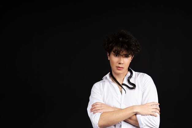 Young handsome man in a white shirt sitting on a chair with a black snake crawling around his neck Isolated on black background