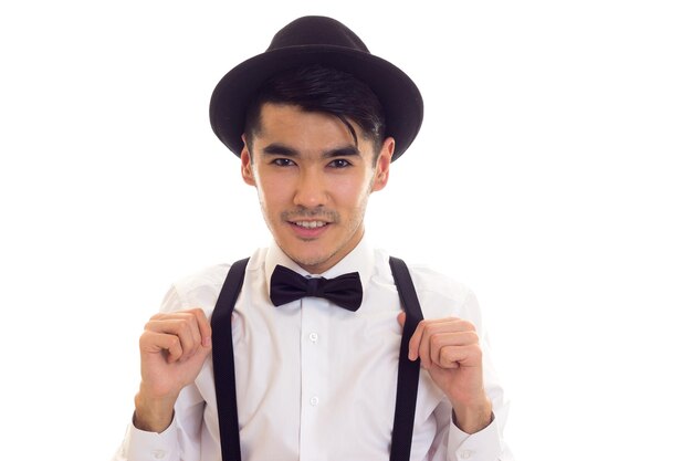 Young handsome man in white shirt with black bow-tie, suspenders and hat on white background in studio