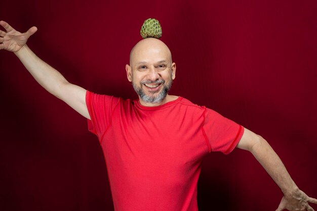 Young handsome man with beard bald head and red shirt standing with pinecone fruit balanced on head against red background positive and healthy person