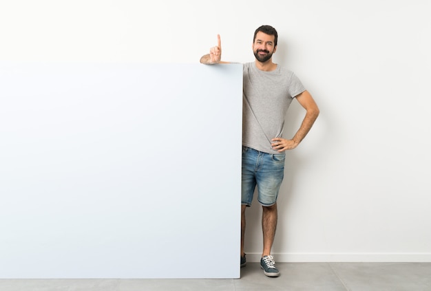 Young handsome man with beard holding a big blue empty placard showing and lifting a finger in sign of the best