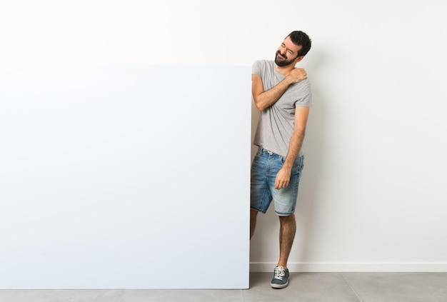 Foto giovane uomo bello con la barba che tiene un grande cartello vuoto blu che soffre di dolore alla spalla per aver fatto uno sforzo