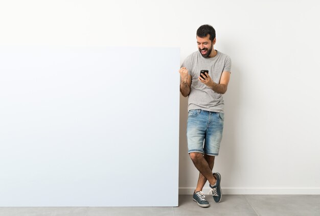 Young handsome man with beard holding a big blue empty placard with phone in victory position