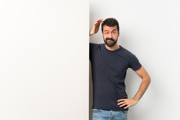 Young handsome man with beard holding a big empty placard having doubts while scratching head