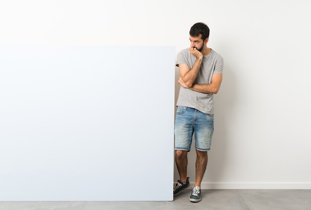 Young handsome man with beard holding a big empty placard having doubts