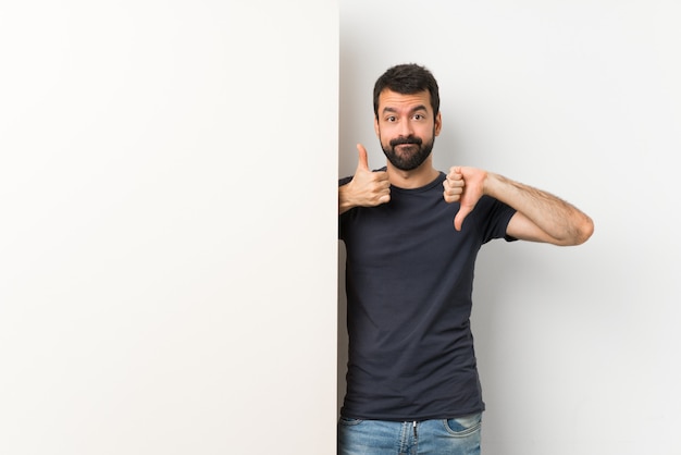 Giovane uomo bello con la barba che tiene un grande cartello vuoto che fa segno buono-cattivo. indeciso tra sì o no