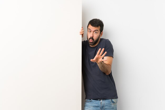 Foto giovane uomo bello con la barba che tiene un grande cartello vuoto nervoso allungando le mani in avanti