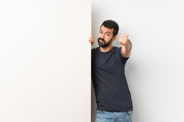 Giovane uomo bello con la barba che tiene un grande cartello vuoto che mostra pollice giù con l'espressione negativa