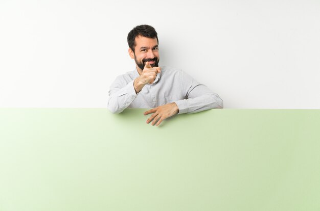 Young handsome man with beard holding a big green empty placard points finger at you with a confident expression