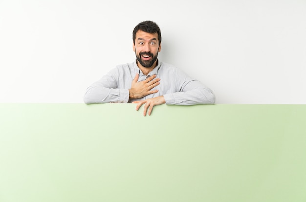 Young handsome man with beard holding a placard surprised and shocked while looking right