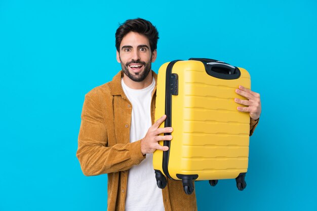 Young handsome man with beard over isolated blue wall in vacation with travel suitcase and surprised