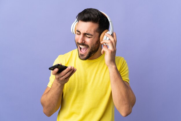 Giovane uomo bello con la barba isolata sulla musica d'ascolto viola con un cellulare e cantando