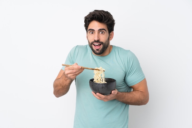 Young handsome man with beard over isolated white wall holding a bowl of noodles with chopsticks and eating it