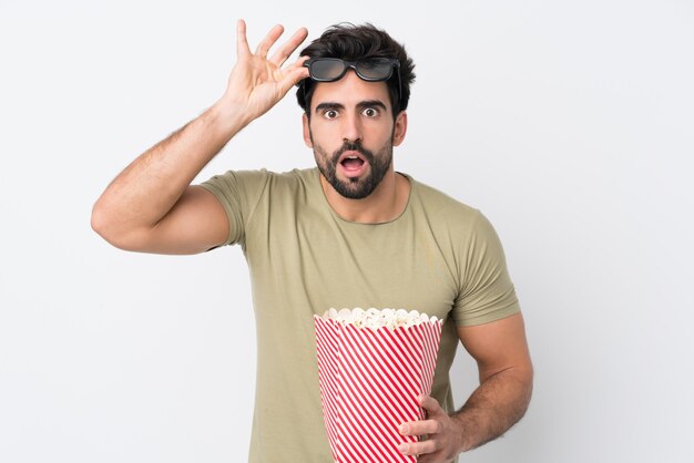 Young handsome man with beard over isolated white wall surprised with 3d glasses and holding a big bucket of popcorns