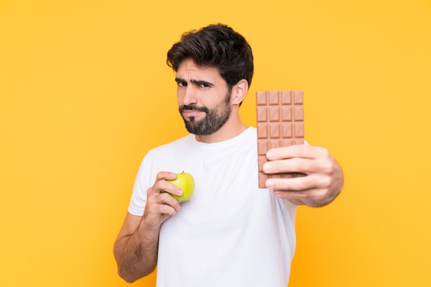 Young handsome man with beard over isolated yellow wall taking a chocolate tablet in one hand and an apple in the other