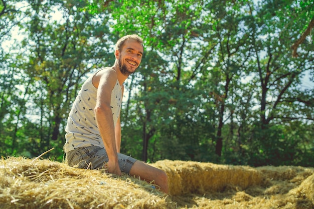 A young handsome man with beard of slim build sits in the hay and holds hay in his hands against the background of green trees in the park