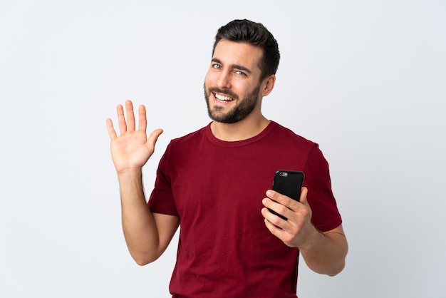 Young handsome man with beard using mobile phone isolated on white saluting with hand with happy expression