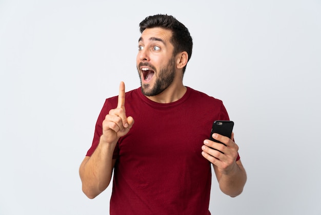 Young handsome man with beard using mobile phone isolated on white wall intending to realizes the solution while lifting a finger up