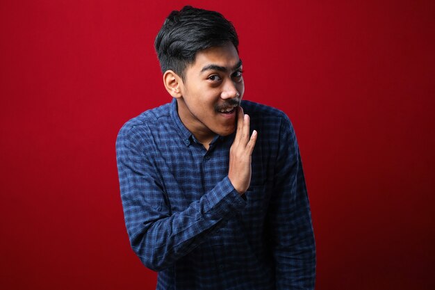 Young handsome man with beard wearing casual shirt over red background hand on mouth telling secret rumor, whispering malicious talk conversation