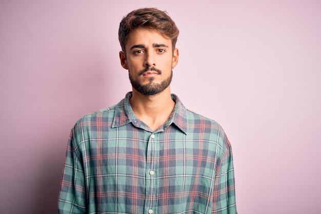 Young handsome man with beard wearing casual shirt standing over pink background Relaxed with serious expression on face Simple and natural looking at the camera