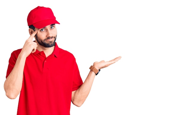 Young handsome man with beard wearing delivery uniform confused and annoyed with open palm showing copy space and pointing finger to forehead think about it