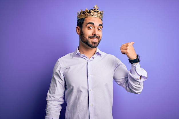 Young handsome man with beard wearing golden crown of king over purple background smiling with happy face looking and pointing to the side with thumb up